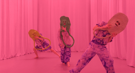 Video still from What Do Stones Smell Like in the Forest: The chorus hold their papier maché masks in front of their faces and face the camera on a pink stage in front of a pink curtain.